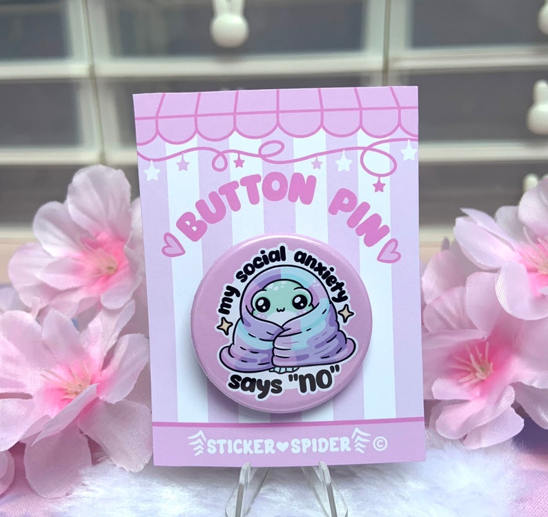 May include: A pink button pin with a cartoon alien wrapped in a purple and blue blanket. The text on the pin reads "My social anxiety says "no"".