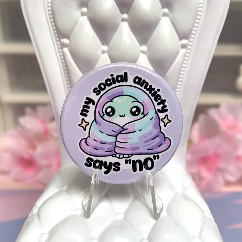May include: A purple button with a cartoon frog wrapped in a blanket. The text on the button reads "My social anxiety says "no"".