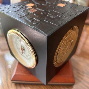 May include: A black cube-shaped decorative clock with a textured top and gold accents. The clock features a barometer and a calendar dial. It sits on a wooden base, showcasing a blend of functionality and design.