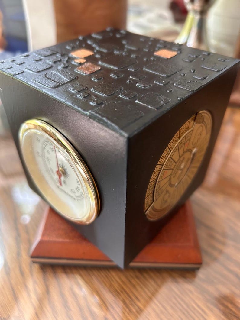 May include: A black cube-shaped decorative clock with a textured top and gold accents. The clock features a barometer and a calendar dial. It sits on a wooden base, showcasing a blend of functionality and design.