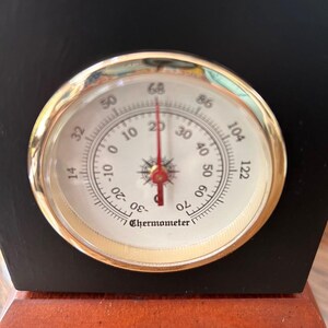 May include: A black and brown wooden thermometer with a gold-colored bezel. The thermometer face is white with black numbers and a red needle. The temperature reads approximately 68 degrees Fahrenheit.