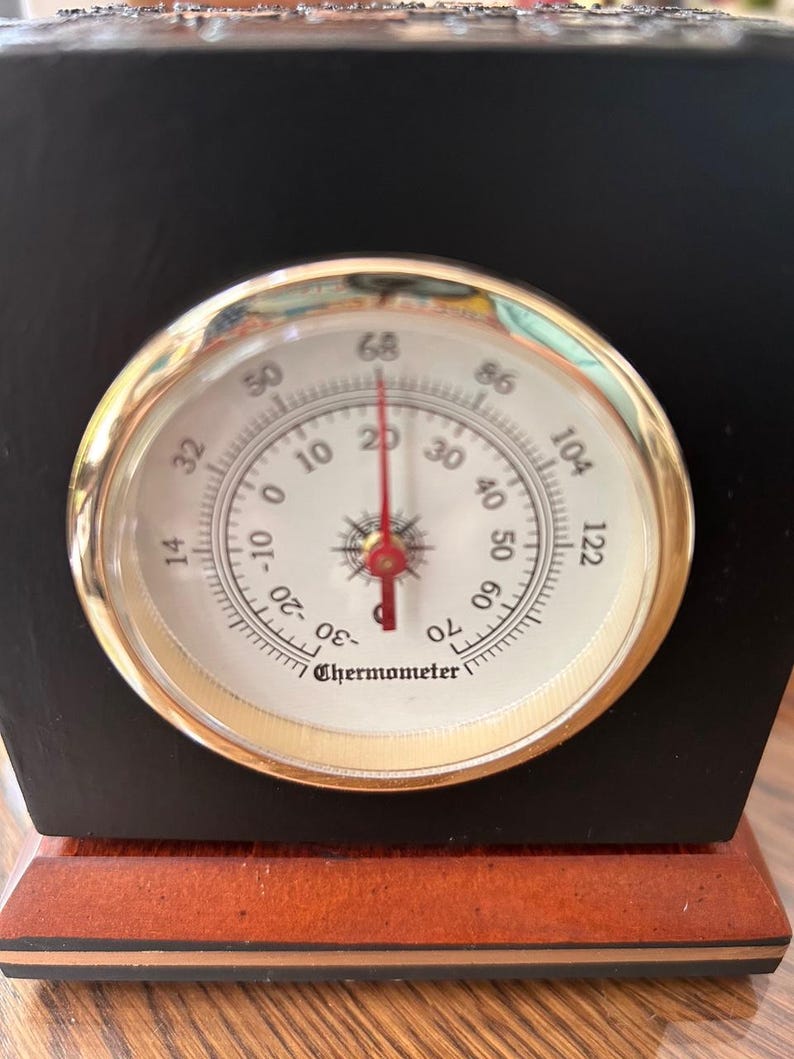 May include: A black and brown wooden thermometer with a gold-colored bezel. The thermometer face is white with black numbers and a red needle. The temperature reads approximately 68 degrees Fahrenheit.