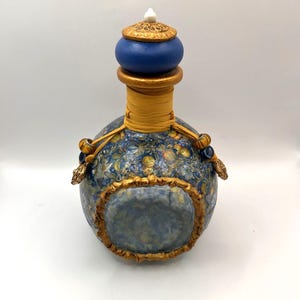 May include: A decorative bottle with a blue and gold design. The bottle has a round shape with a blue stopper and gold accents. The body of the bottle is decorated with a floral pattern in shades of blue, yellow, and green.