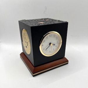 May include: A black cube clock with a gold-rimmed clock face and a gold-colored dial on the side. The clock sits on a wooden base with a dark brown top and a gold accent. The clock's face shows the time as 7:40.