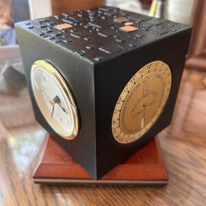 May include: A black cube-shaped clock with a gold-rimmed clock face and a gold inch/metric converter dial. The cube sits on a wooden base. The top of the cube has a textured design with copper-colored accents.