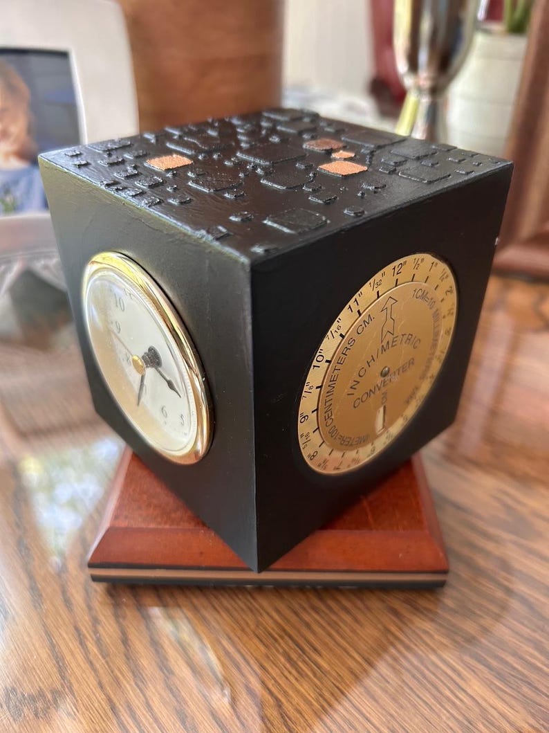 May include: A black cube-shaped clock with a gold-rimmed clock face and a gold inch/metric converter dial. The cube sits on a wooden base. The top of the cube has a textured design with copper-colored accents.