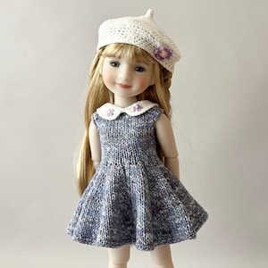 Peut inclure: Une poupée vêtue d'une robe bleue tricotée avec un col blanc et un béret blanc assorti. Le béret est orné d'une petite fleur violette. La poupée a de longs cheveux blonds et sourit.