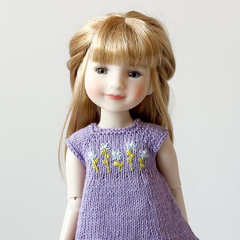 May include: A doll with blonde hair wearing a lavender knitted dress. The dress is decorated with embroidered white and yellow flowers. The doll has a calm expression and is set against a plain background.