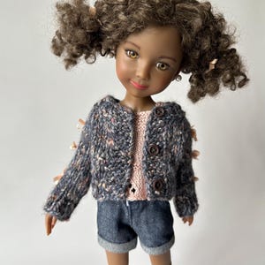 May include: A doll with brown curly hair wearing a blue and pink knit cardigan with wooden buttons, a pink top, and denim shorts. The doll has brown eyes and is wearing small pearl earrings.
