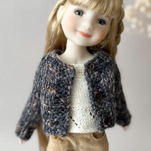 Peut inclure: Une poupée vêtue d'une veste en tricot bleu foncé moucheté avec des boutons marron, sur un pull crème. La poupée a de longs cheveux blonds et porte un pantalon beige. La veste est courte.