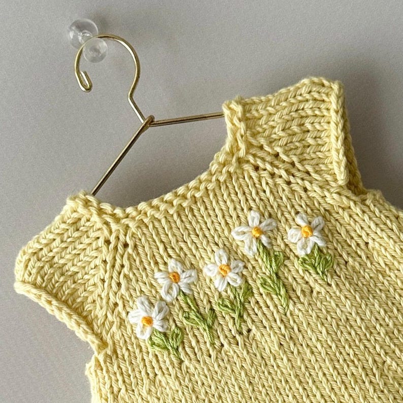 May include: A yellow knitted baby dress with white and yellow daisy embroidery. The sleeveless dress is hanging on a gold hanger. The daisies have green stems and leaves.