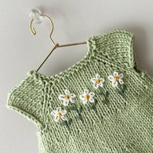 May include: A pale green knitted top with a daisy floral design. The sleeveless garment has white daisies with yellow centres and green stems embroidered on the front. It hangs on a gold-coloured hanger.