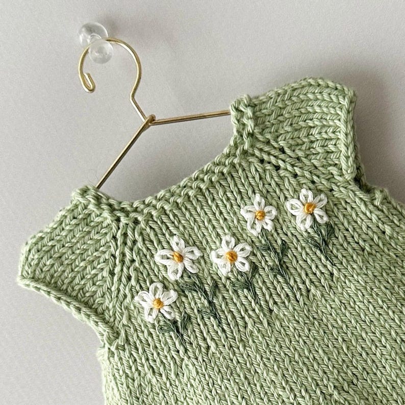 May include: A pale green knitted top with a daisy floral design. The sleeveless garment has white daisies with yellow centres and green stems embroidered on the front. It hangs on a gold-coloured hanger.