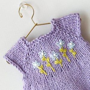 May include: A lilac knitted garment, possibly a baby dress or top, decorated with embroidered pale blue flowers and green stems. The item is displayed on a gold-coloured hanger against a white background. The knitwear has a ribbed neckline and a textured stitch.