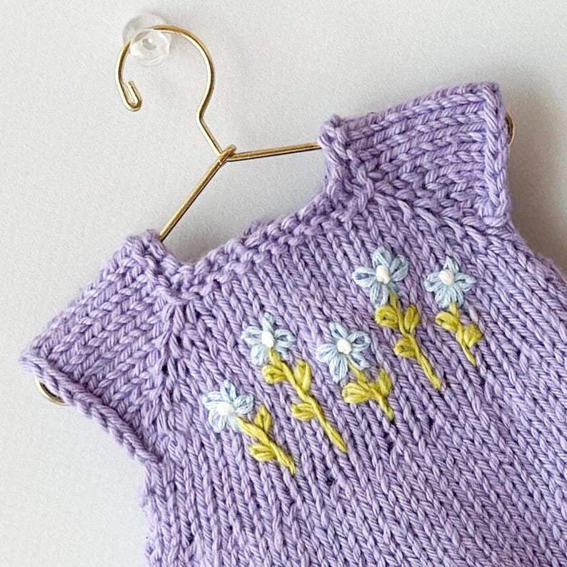 May include: A lilac knitted garment, possibly a baby dress or top, decorated with embroidered pale blue flowers and green stems. The item is displayed on a gold-coloured hanger against a white background. The knitwear has a ribbed neckline and a textured stitch.