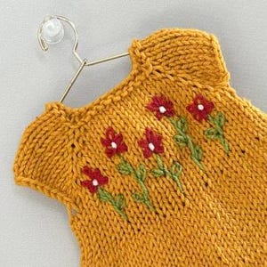 May include: Mustard yellow knitted top with embroidered red flowers and green stems. The top is displayed on a gold-coloured hanger. The floral design adds a touch of handmade charm to this garment.