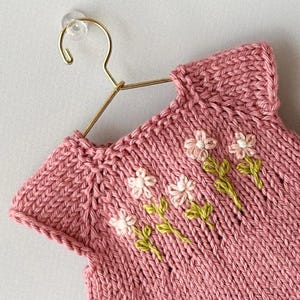 May include: A pink knitted garment with a floral design. The sleeveless top is displayed on a gold hanger. The flowers are white with green stems, embroidered on the front of the garment. The knitwear has a ribbed texture.