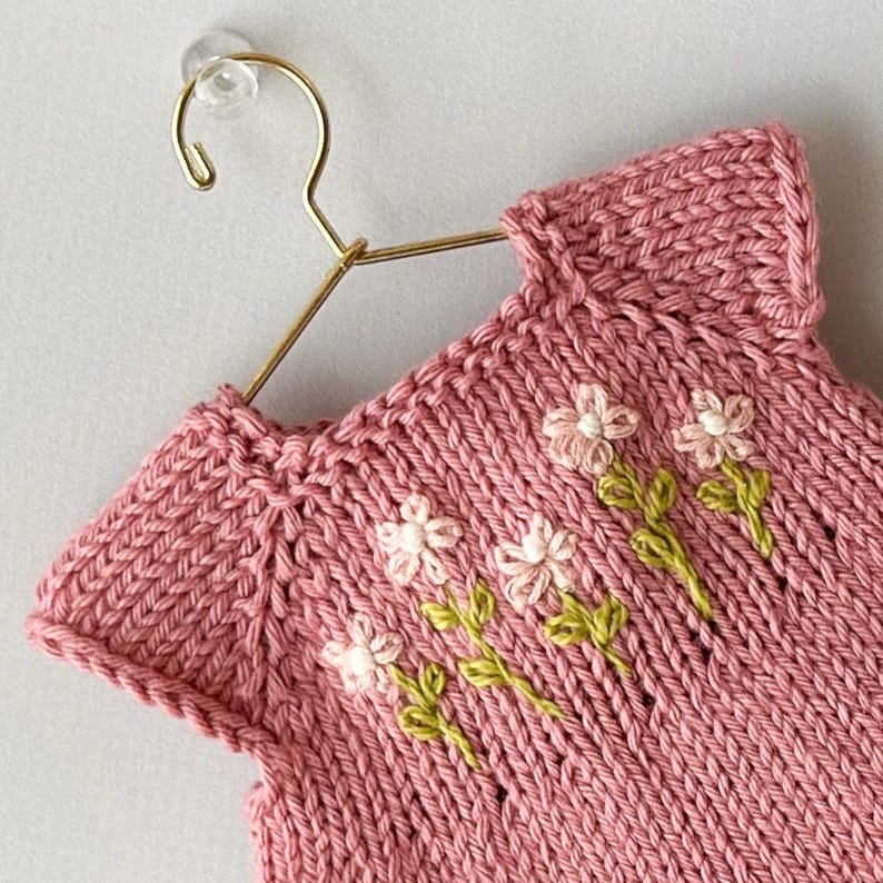 May include: A pink knitted garment with a floral design. The sleeveless top is displayed on a gold hanger. The flowers are white with green stems, embroidered on the front of the garment. The knitwear has a ribbed texture.