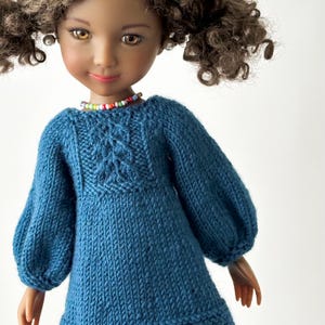 May include: A doll in a blue knitted dress featuring a cable knit pattern on the bodice and long, puffed sleeves. The doll has brown curly hair and a colourful beaded necklace. The doll's skin tone is light brown.