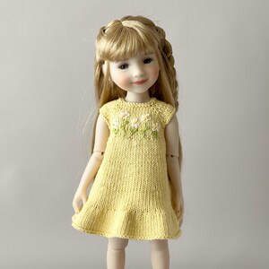 May include: A doll with long blonde hair wearing a yellow knitted dress. The dress has a ruffle hem and a floral design of white daisies with green leaves embroidered on the bodice. The doll has a serene expression.