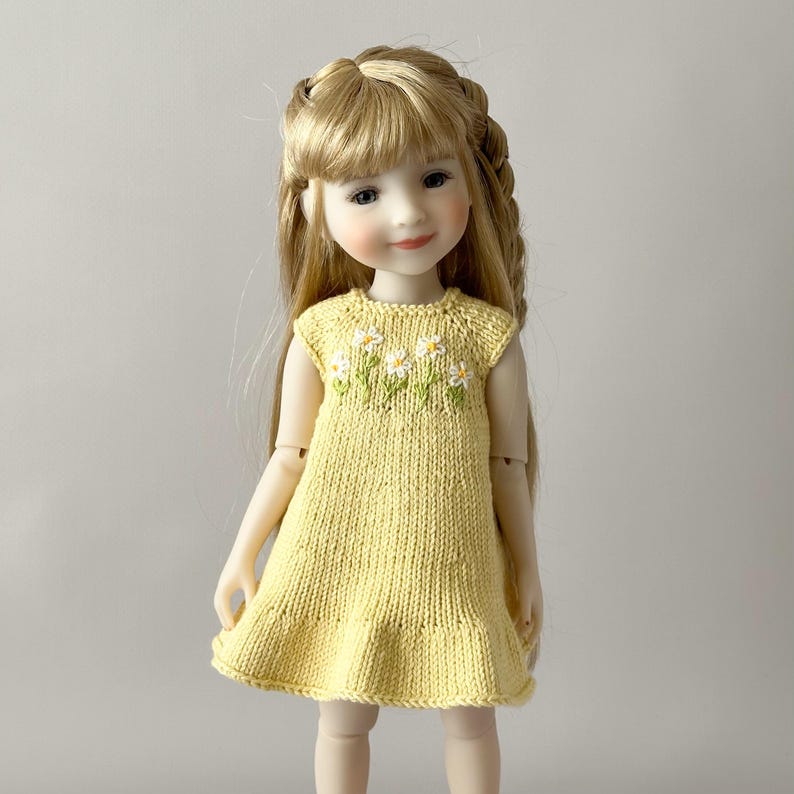 May include: A doll with long blonde hair wearing a yellow knitted dress. The dress has a ruffle hem and a floral design of white daisies with green leaves embroidered on the bodice. The doll has a serene expression.