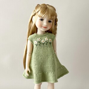 May include: A doll in a light green knitted dress, adorned with white daisy embroidery on the bodice. The doll has long blonde hair styled with braids. The dress features a flared skirt and cap sleeves.