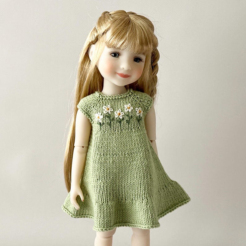 May include: A doll in a light green knitted dress, adorned with white daisy embroidery on the bodice. The doll has long blonde hair styled with braids. The dress features a flared skirt and cap sleeves.