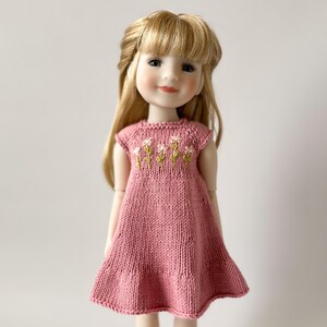 May include: A doll with long blonde hair wearing a pink knitted dress. The dress has a floral embroidery design on the bodice. The doll has a gentle expression and is set against a plain background.