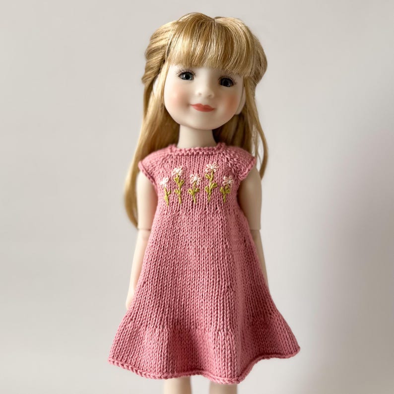 May include: A doll with long blonde hair wearing a pink knitted dress. The dress has a floral embroidery design on the bodice. The doll has a gentle expression and is set against a plain background.