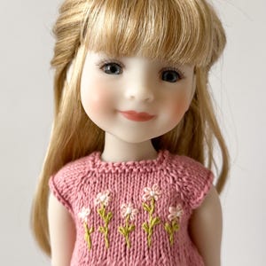 May include: A doll with blonde hair and blue eyes wearing a pink knitted dress with embroidered white flowers and green stems. The doll has rosy cheeks and a gentle smile.