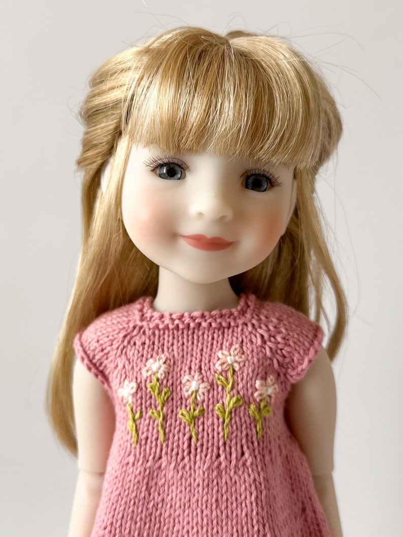 May include: A doll with blonde hair and blue eyes wearing a pink knitted dress with embroidered white flowers and green stems. The doll has rosy cheeks and a gentle smile.