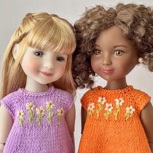 May include: Two dolls with detailed faces and hair. One doll has blonde hair and wears a lilac knit dress with daisy embroidery. The other doll has brown curly hair and wears an orange knit dress with daisy embroidery.
