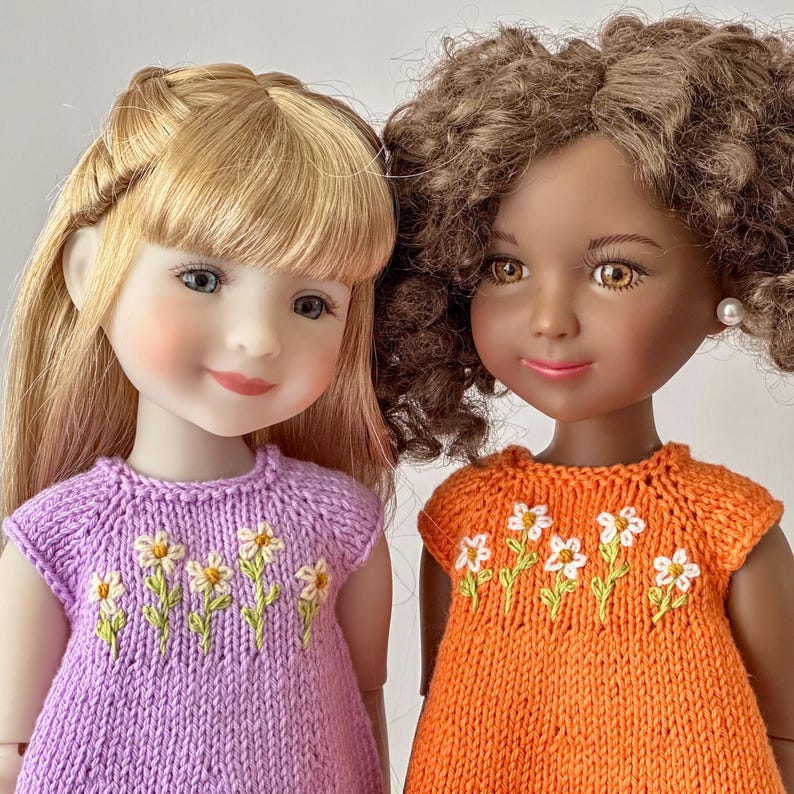 May include: Two dolls with detailed faces and hair. One doll has blonde hair and wears a lilac knit dress with daisy embroidery. The other doll has brown curly hair and wears an orange knit dress with daisy embroidery.