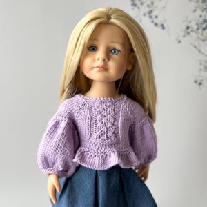 Peut inclure: Une poupée portant un pull en tricot lavande avec un ourlet à volants et des manches longues. La poupée a de longs cheveux blonds et des yeux bleus.
