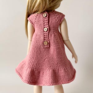 May include: A dusty rose knitted doll dress with a flared skirt and cap sleeves. The back of the dress has three wooden buttons. The dress is made of a soft, textured knit fabric.