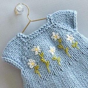 May include: A pale blue knitted baby dress with embroidered white daisies and green stems. The dress is displayed on a gold-coloured hanger. The knitwear features a round neckline and short sleeves.