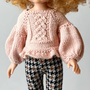 Puede incluir: Un suéter de punto rosa con un patrón de trenza y mangas abullonadas. El suéter tiene un dobladillo con volantes y lo lleva una muñeca.
