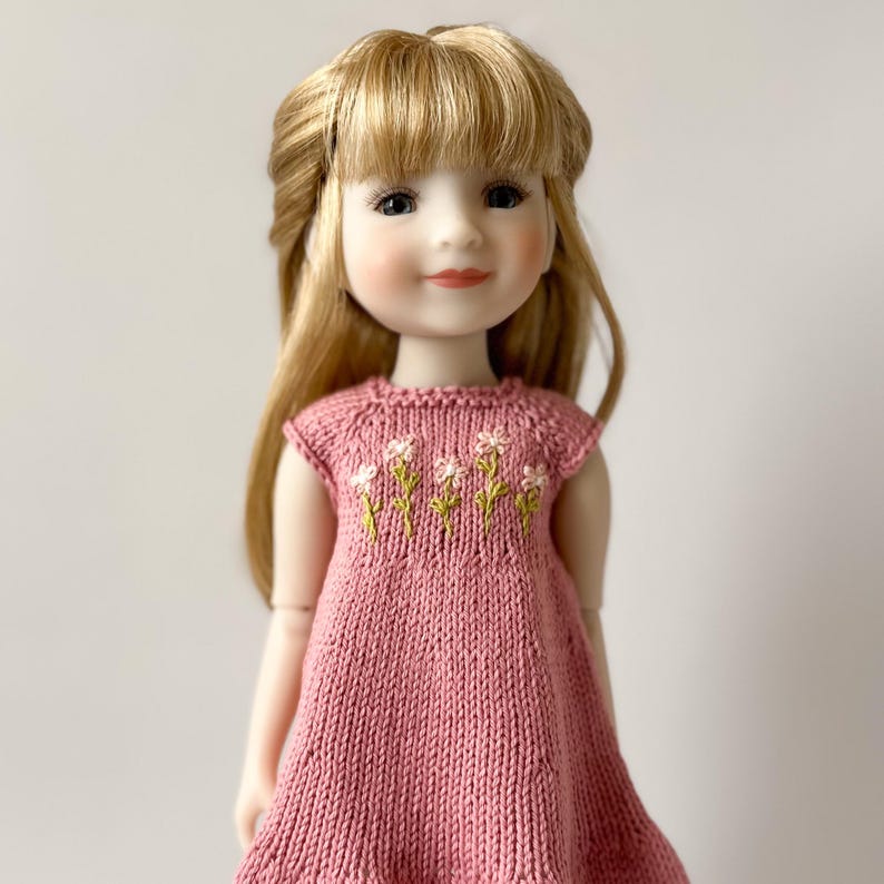 May include: A doll with blonde hair and a pink knitted dress. The dress has embroidered white and yellow flowers on the bodice. The doll has a calm expression and is finely detailed.
