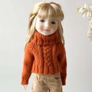 Op de afbeelding: Een pop met een oranje kabelgebreide coltrui en een beige corduroy broek. De pop heeft lang blond haar en glimlacht. De trui heeft een textuurontwerp en een geribbelde kraag. Witte achtergrond.