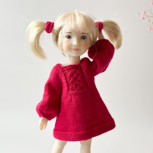 May include: A doll wearing a red knitted dress with a cable knit pattern on the front. The doll has blonde hair in pigtails and blue eyes.