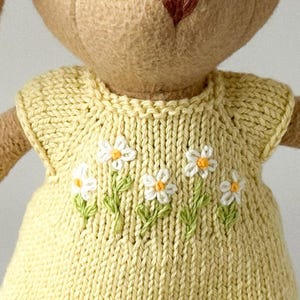May include: A close-up of a knitted yellow dress with white and yellow daisy embroidery. The dress has a round neckline and short sleeves. The dress is made of a soft, textured yarn.