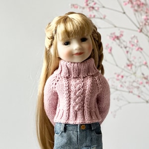 May include: A doll with long blonde hair and a pink turtleneck jumper with a cable knit design. The doll is wearing denim jeans. The background is a soft white with pink flowers.