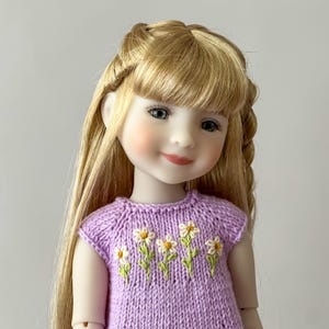 May include: A doll with long blonde hair and blue eyes, wearing a lilac knit dress embroidered with white and yellow daisies. The doll has rosy cheeks and a gentle smile, with a braided hairstyle.