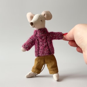 May include: A small, white stuffed mouse wearing a pink knitted jumper and brown trousers. The mouse is standing on a white surface.