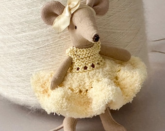 May include: A beige fabric mouse doll featuring a cream-colored satin ribbon bow on its head. The mouse wears a pale yellow knitted bodice paired with a fluffy, textured, light yellow skirt. The doll has stitched facial features.
