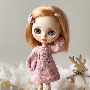 May include: A pink knitted dress with a cable knit pattern and long sleeves. The dress is worn by a doll with blonde hair and blue eyes.