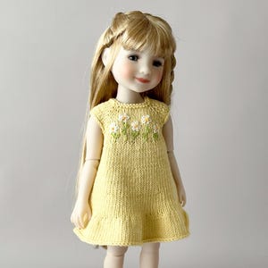 May include: A doll with blonde hair wearing a yellow knitted dress with white and green daisy embroidery. The dress has a ruffled hem and sleeveless design. The doll has rosy cheeks and is posed against a neutral background.