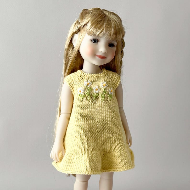 May include: A doll with blonde hair wearing a yellow knitted dress with white and green daisy embroidery. The dress has a ruffled hem and sleeveless design. The doll has rosy cheeks and is posed against a neutral background.