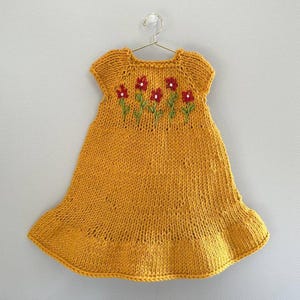 May include: Mustard yellow knitted dress with short sleeves and a flared skirt, hanging on a metal hanger. The dress features embroidered red flowers with green stems across the chest. A charming handmade garment.