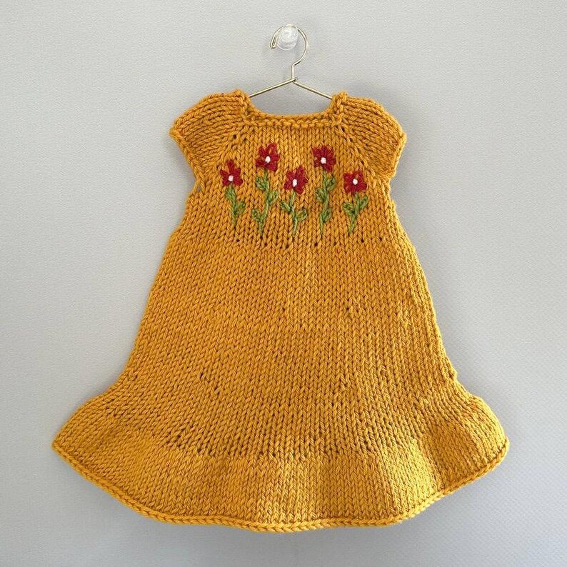May include: Mustard yellow knitted dress with short sleeves and a flared skirt, hanging on a metal hanger. The dress features embroidered red flowers with green stems across the chest. A charming handmade garment.